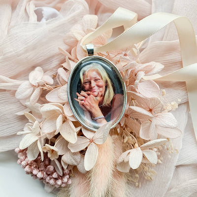 A memorial pendant or memorial charm in the design called Modern Oval, showing a photo of a mother or grandmother. The pendant is nestled on soft peach fabric and dried hydrangeas and is intended to be tied to a wedding bouquet with ribbon or hung from a chain to wear, to include a deceased relative on your wedding day.