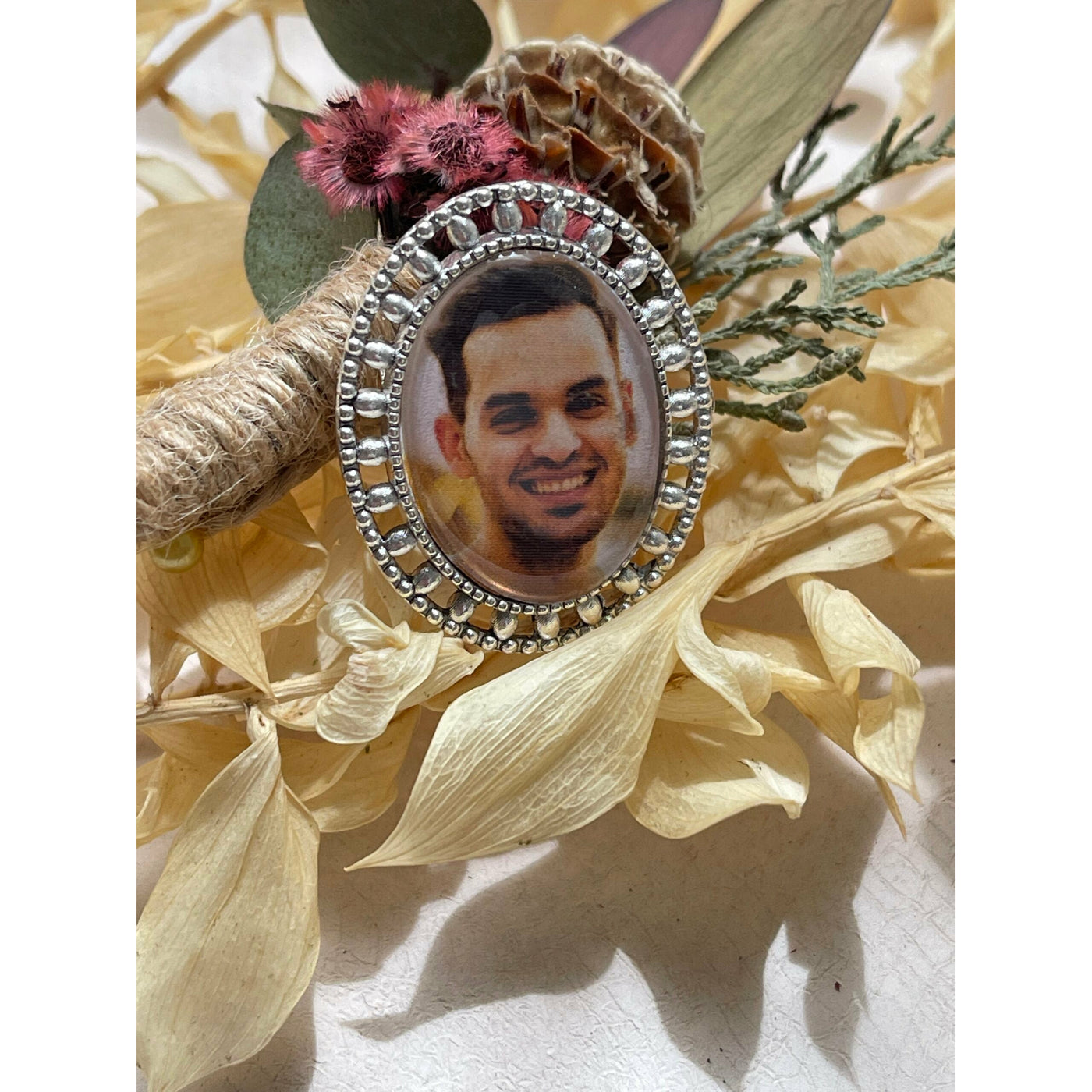 A memorial brooch or memorial pin in the design called Brogue, showing a photo of a young man. The pin is nestled on dried Australian native flowers and is intended to be pinned to a wedding bouquet or wedding jacket to include a deceased relative on your wedding day.