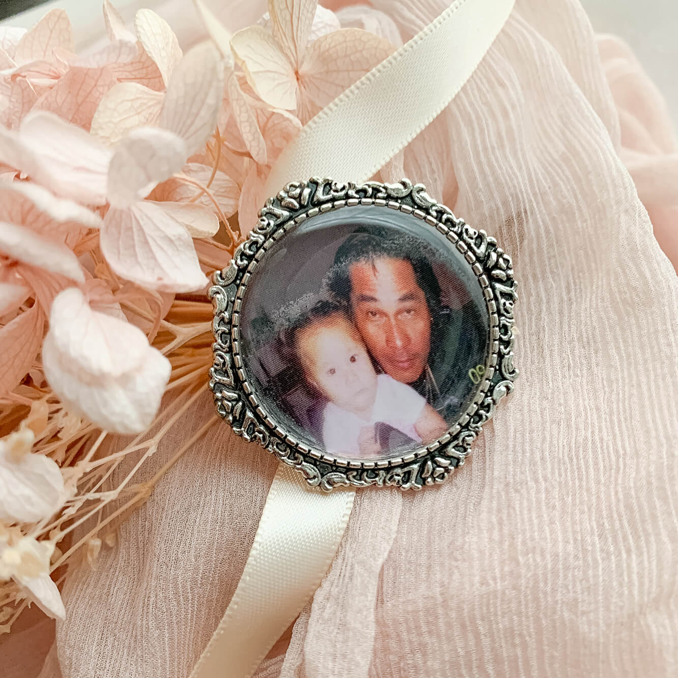 A memorial brooch or memorial pin in the design called Lace, showing a photo of a father and his child. The pin is nestled on soft peach fabric and dried hydrangeas and is intended to be pinned to a wedding bouquet or wedding jacket to include a deceased relative on your wedding day.