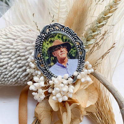 A memorial pin or memorial brooch in the design called Amore, showing a photo of a grandfather inside. The pendant is nestled on neutral textured surface and dried gold flowers and is intended to be pinned to a wedding bouquet or suit jacket, to include a deceased relative on your wedding day.