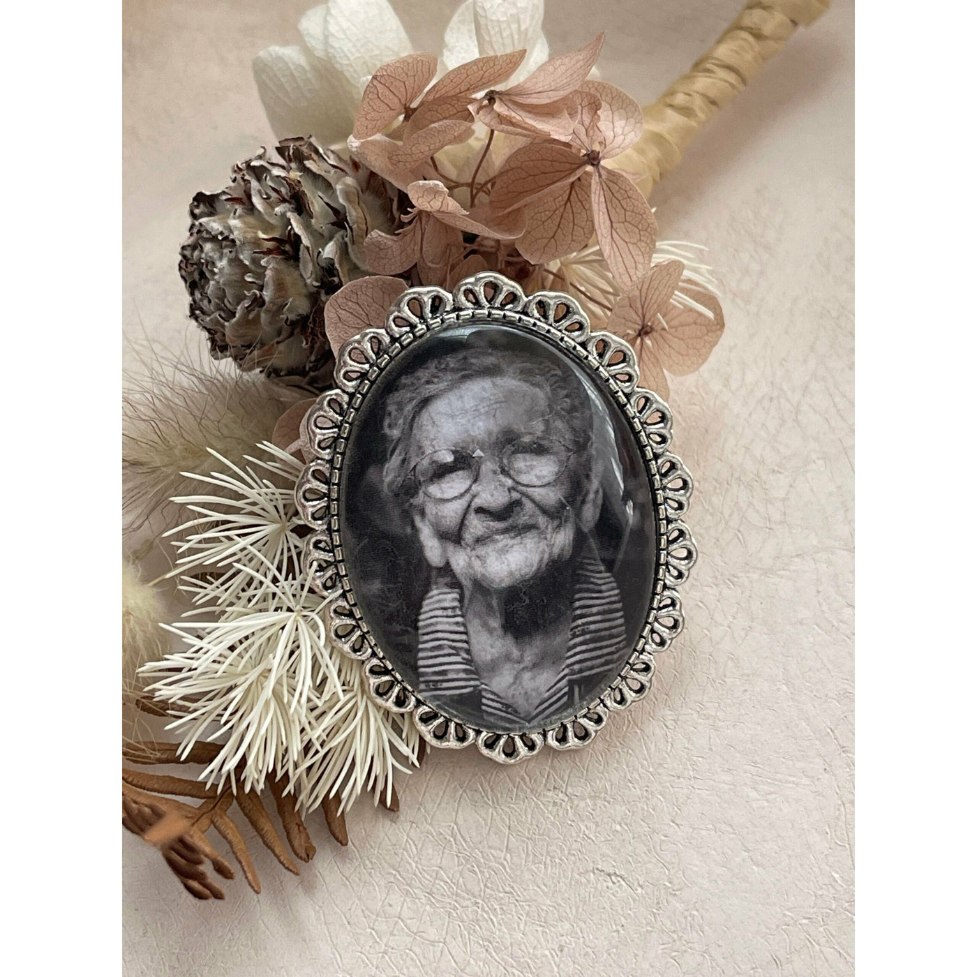 An oval memorial pin or brooch that is no longer manufactured or available to purchase. This memorial pin shows an old woman in black and white and was made to be pinned on to a wedding bouquet or wedding jacket to honour a deceased loved one on your wedding day.