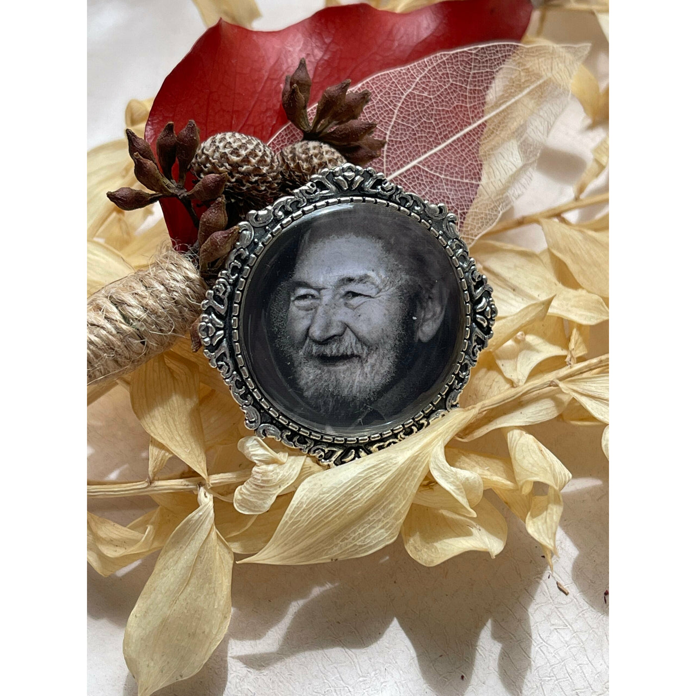 A memorial brooch or memorial pin in the design called Lace showing a photo of a grandfather. The pin is attached to a boutonniere of dried Australian native flowers and is intended to be pinned to a wedding bouquet or wedding jacket to include a deceased relative on your wedding day.