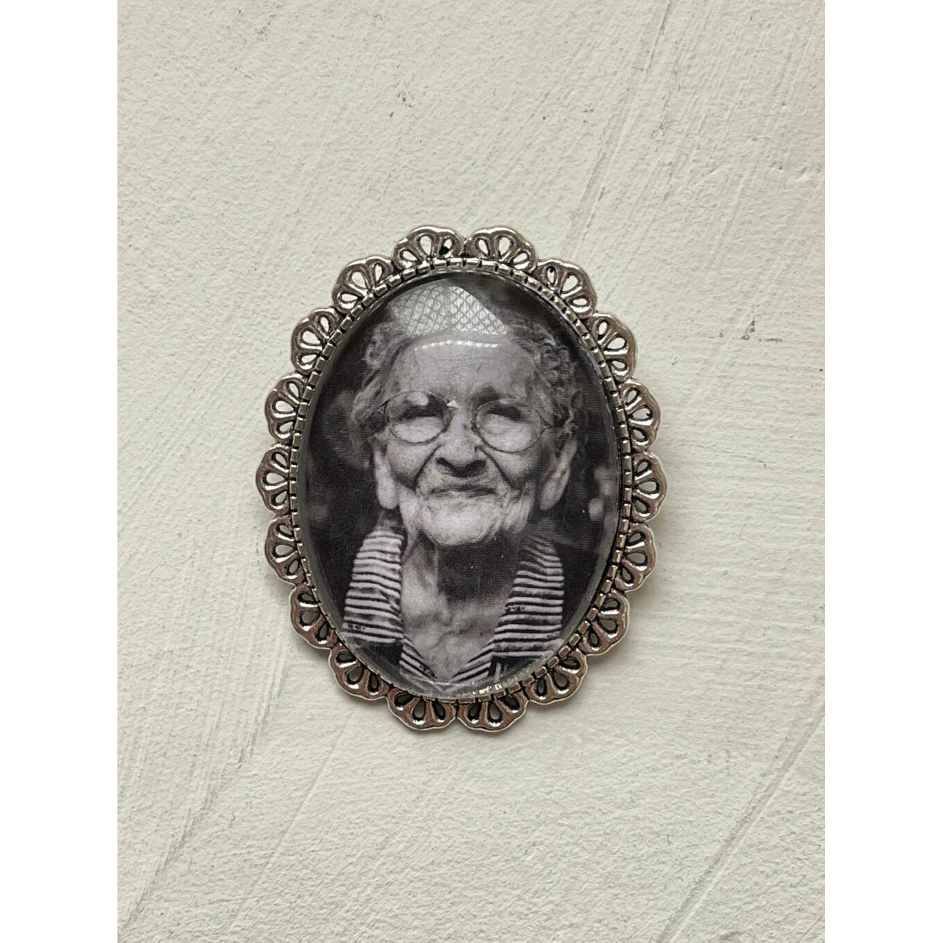 An oval memorial pin or brooch that is no longer manufactured or available to purchase. This memorial pin shows an old woman in black and white and was made to be pinned on to a wedding bouquet or wedding jacket to honour a deceased loved one on your wedding day.