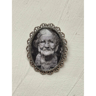 An oval memorial pin or brooch that is no longer manufactured or available to purchase. This memorial pin shows an old woman in black and white and was made to be pinned on to a wedding bouquet or wedding jacket to honour a deceased loved one on your wedding day.