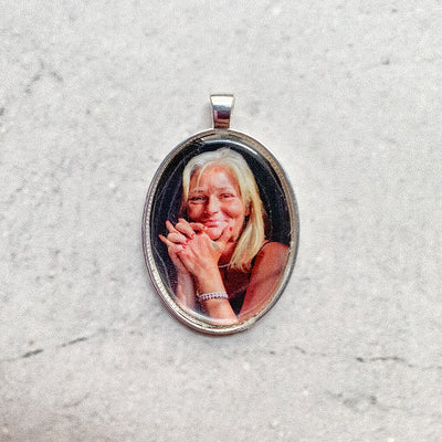 A memorial pendant or memorial charm in the design called Modern Oval, showing a photo of a mother or grandmother. The pendant is nestled on a neutral white textured background, and is intended to be tied to a wedding bouquet with ribbon or hung from a chain to wear, to include a deceased relative on your wedding day.