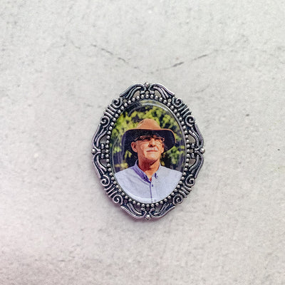 A memorial pin or memorial brooch in the design called Amore, showing a photo of a grandfather inside. The pendant is nestled on neutral white textured background and is intended to be pinned to a wedding bouquet or suit jacket, to include a deceased relative on your wedding day.