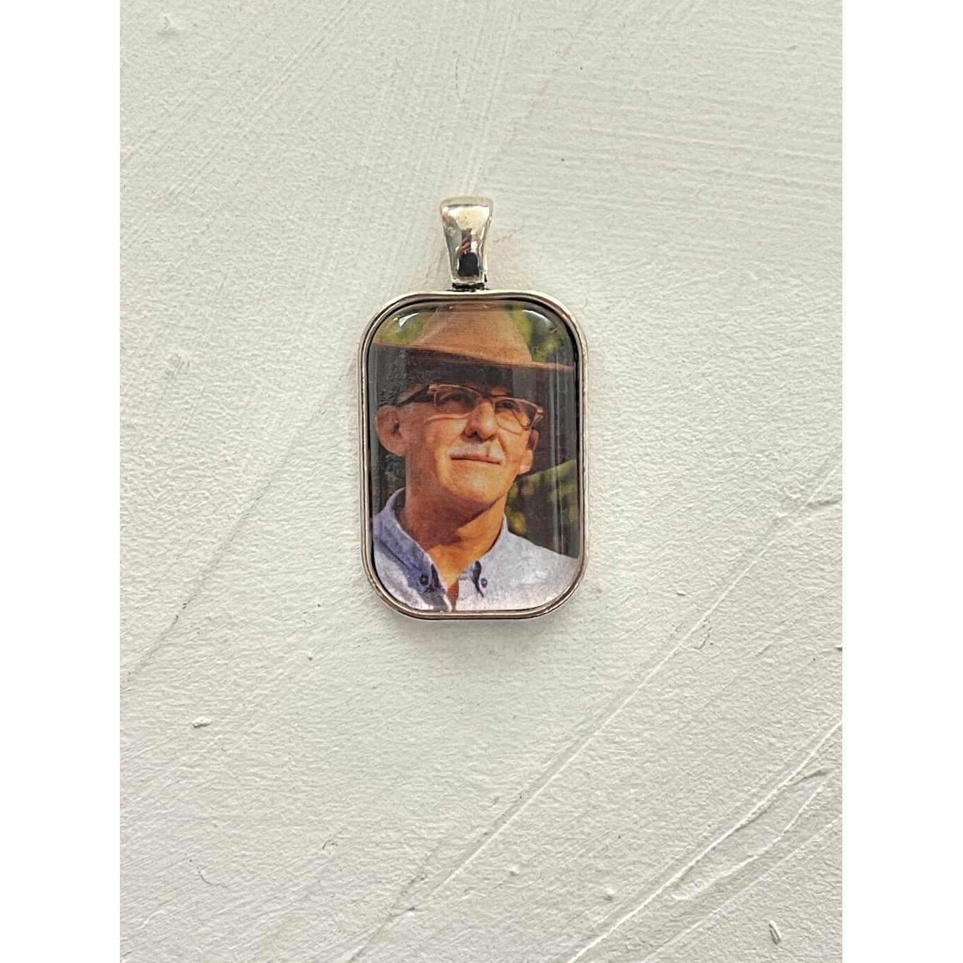A memorial pendant or memorial charm in the design called Rectangle Memorial Photo Pendant, showing a photo of a grandfather inside. The pendant is shown on a white neutral textured background and is intended to be tied to a wedding bouquet with ribbon or hung from a chain to wear, to include a deceased relative on your wedding day.