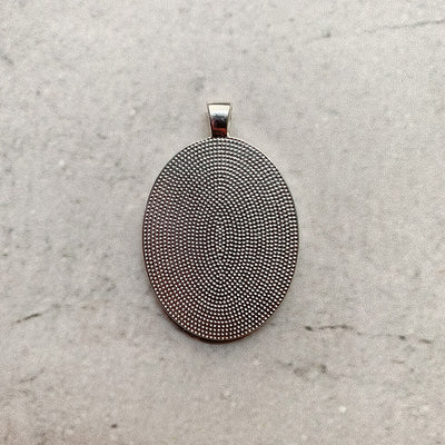 A memorial pendant or memorial charm in the design called Modern Oval, showing the back of the pendant on a neutral white textured background. The pin is nestled on soft peach fabric and dried hydrangeas and is intended to be tied to a wedding bouquet with ribbon or hung from a chain to wear, to include a deceased relative on your wedding day.