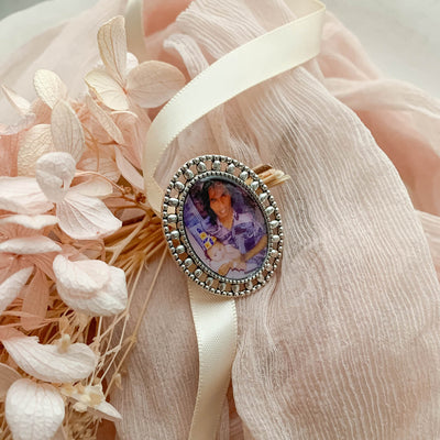 A memorial brooch or memorial pin in the design called Brogue, showing a photo of a young father and his child. The pin is nestled on soft peach fabric and dried hydrangeas and is intended to be pinned to a wedding bouquet or wedding jacket to include a deceased relative on your wedding day.