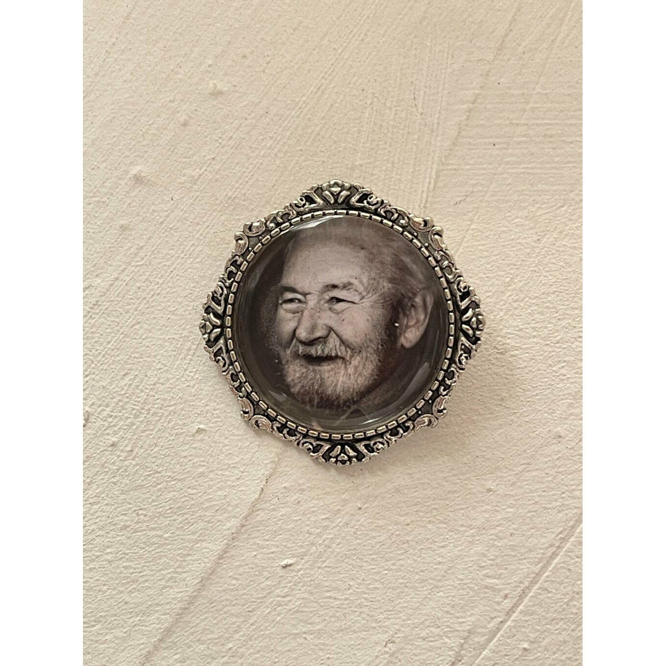 A memorial brooch or memorial pin in the design called Lace showing a photo of a grandfather. The pin is show on a neutral white background and is intended to be pinned to a wedding bouquet or wedding jacket to include a deceased relative on your wedding day.