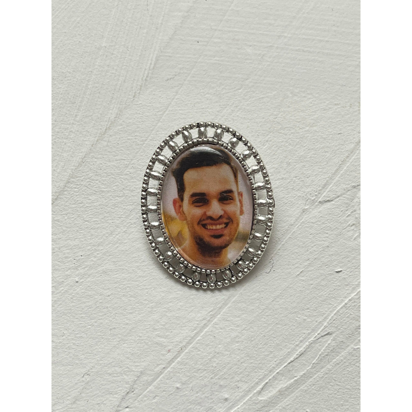 A memorial brooch or memorial pin in the design called Brogue, showing a photo of a young man. The pin is placed on a neutral white textured background and is intended to be pinned to a wedding bouquet or wedding jacket to include a deceased relative on your wedding day.
