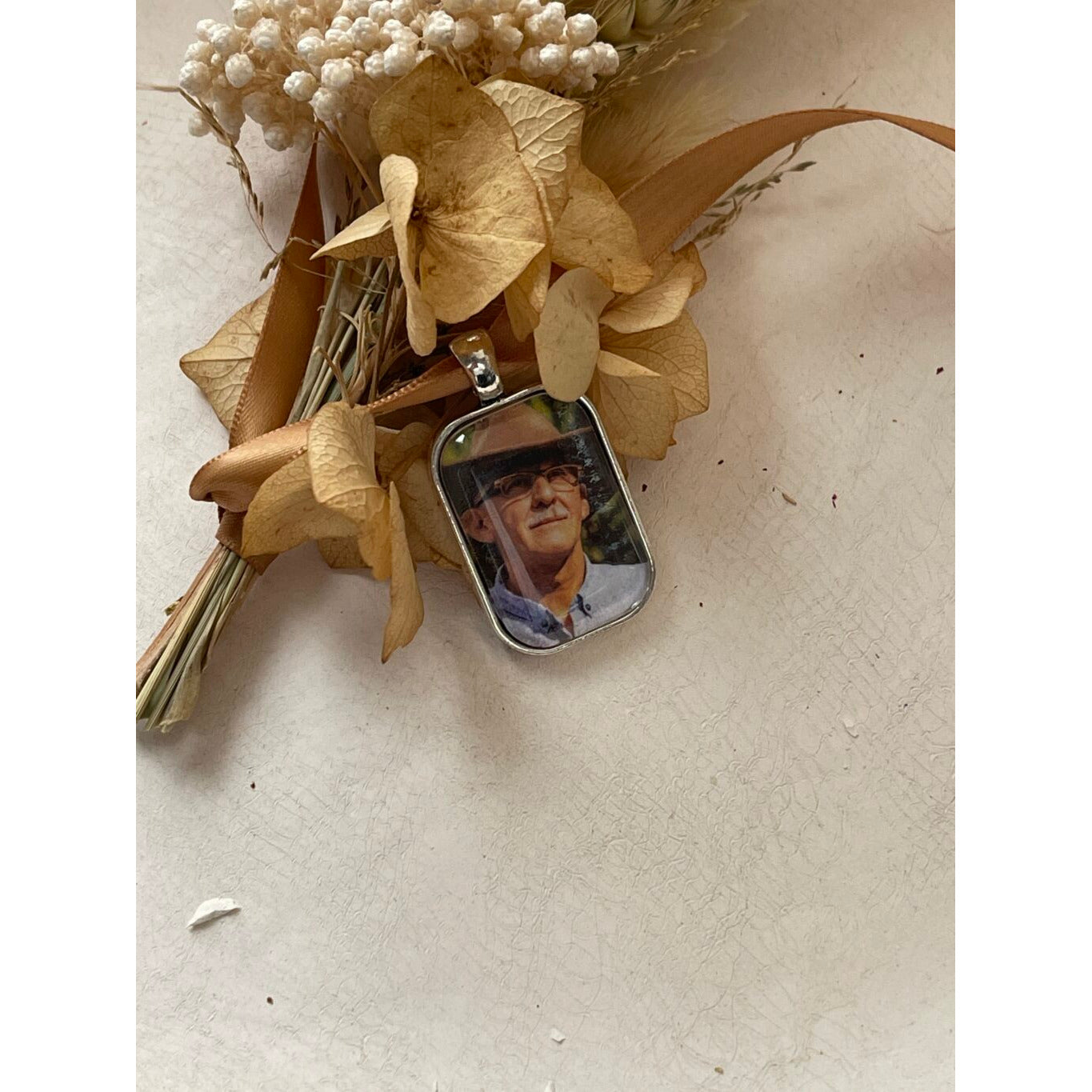 A memorial pendant or memorial charm in the design called Rectangle Memorial Photo Pendant, showing a photo of a grandfather inside. The pendant is nestled on neutral textured surface and dried gold flowers and is intended to be tied to a wedding bouquet with ribbon or hung from a chain to wear, to include a deceased relative on your wedding day.