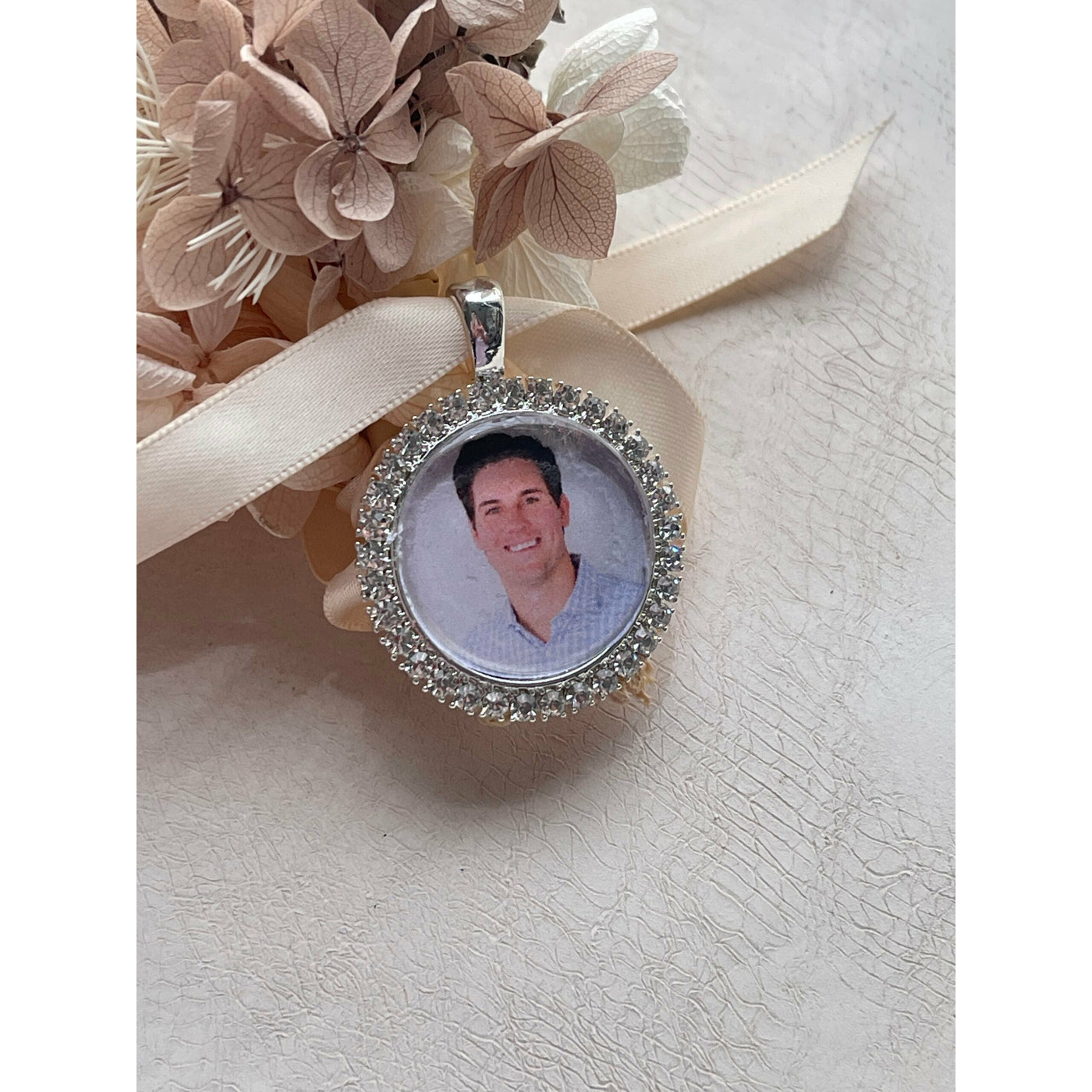 A memorial pendant or memorial charm in a design called Crystal Round, with crystal diamantes around a circular photo with a sturdy silver loop and a photo of a young man. This pendant is designed to be tied on to ribbon around a bouquet or hung from a chain to keep the memory of a deceased loved on with you on your wedding day.